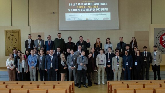 Organizatorzy i prelegenci pozują do wspólnego zdjęcia na tle plakatu konferencji.